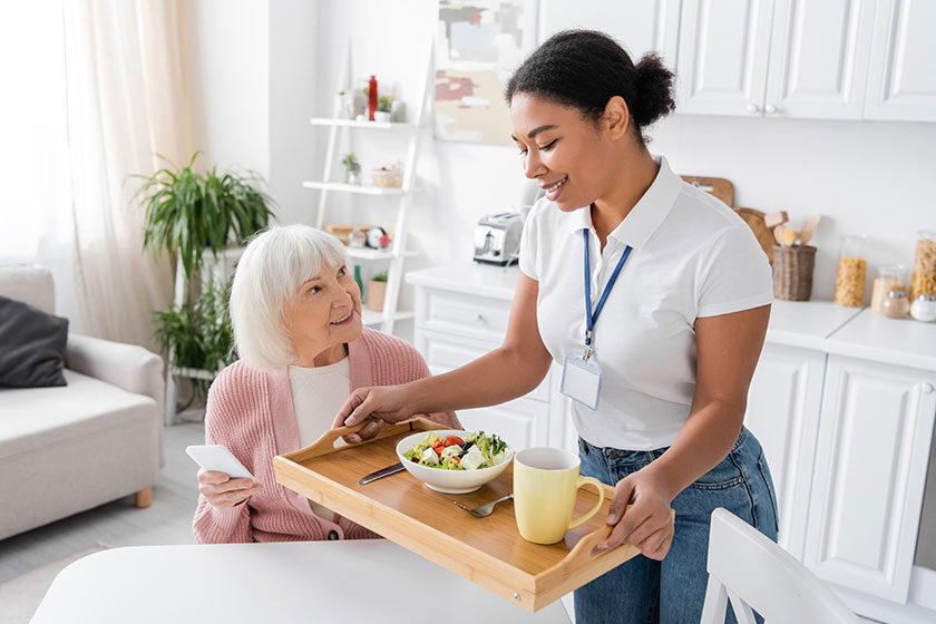 Happy multiracial social worker holding tray with lunch for senior woman with grey hair