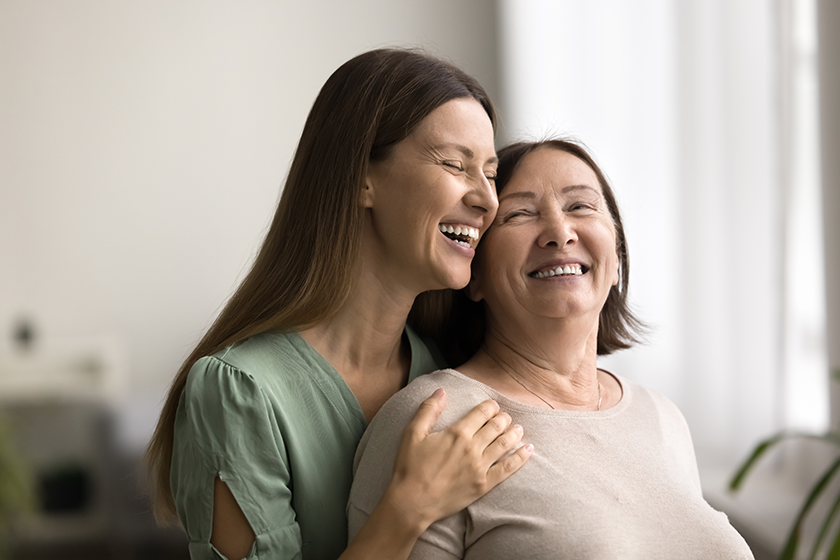 Joyful adult child woman hugging happy mom from behind, holding shoulders, talking, smiling, laughing, enjoying closeness, embracing mother with love, affection, care, gratitude 