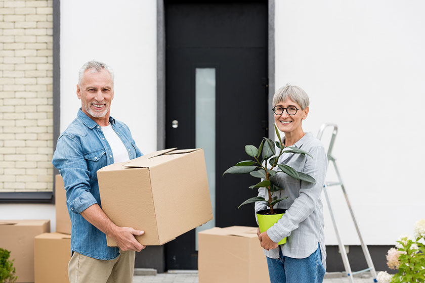 Mature man holding box and smiling woman holding plant near new house