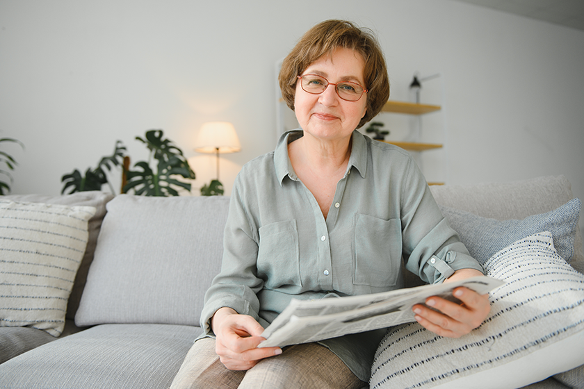 Senior lady reading her newspaper at home relaxing on a couch and peering over the top at the viewer.